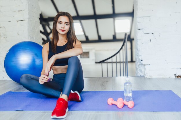 Mujer bonita sentada en su colchoneta con bola azul y pesas mirando a la cámara