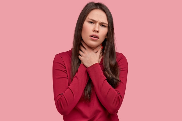 Foto gratuita la mujer bonita se siente mal, mantiene las manos en el cuello, tiene inflamación de la garganta, usa ropa roja, sufre asfixia