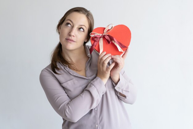 Mujer bonita soñadora que sostiene la caja de regalo en forma de corazón roja