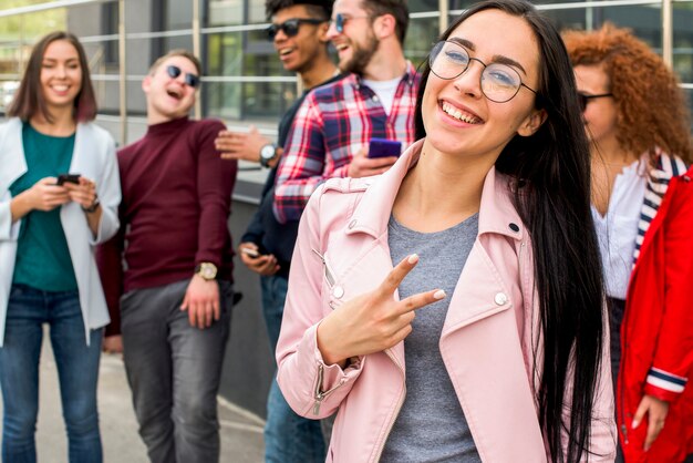 Mujer bonita sonriente que se coloca delante de sus amigos que gesticulan la muestra de la victoria