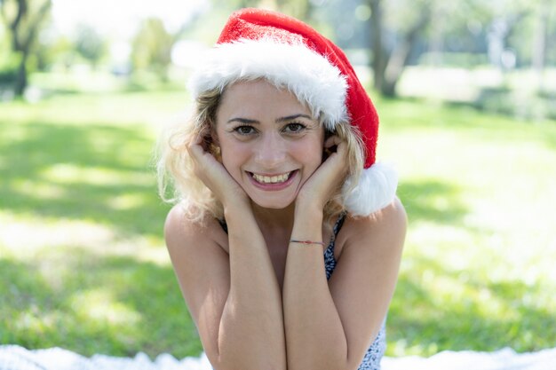 Mujer bonita sonriente que lleva el sombrero de Papá Noel en parque del verano