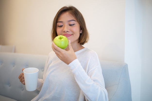 Mujer bonita sonriente que se sienta en el sofá con la taza de la manzana y del té