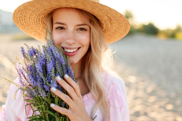 Mujer bonita sonriente con sombrero de paja posando en la playa soleada cerca del océano con ramo de flores. Retrato de cerca.