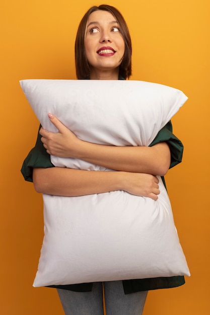 Mujer bonita sonriente sostiene la almohada y mira al lado aislado en la pared naranja