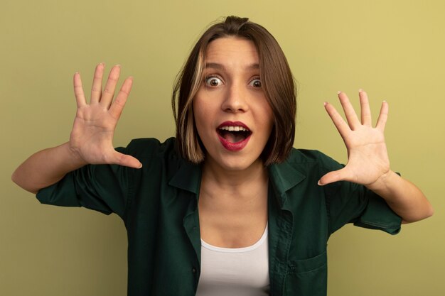 Mujer bonita sorprendida se encuentra con las manos levantadas aisladas en la pared verde oliva