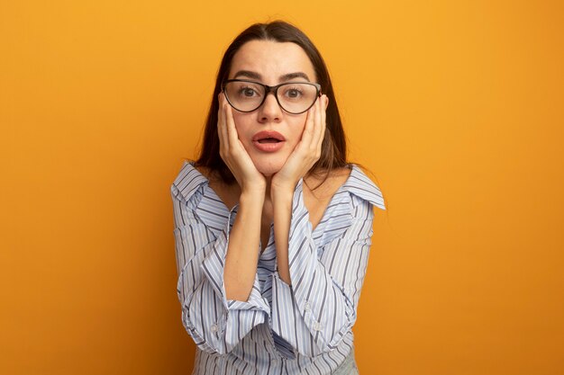 Mujer bonita sorprendida en gafas ópticas pone las manos en la cara y mira al frente aislado en la pared naranja