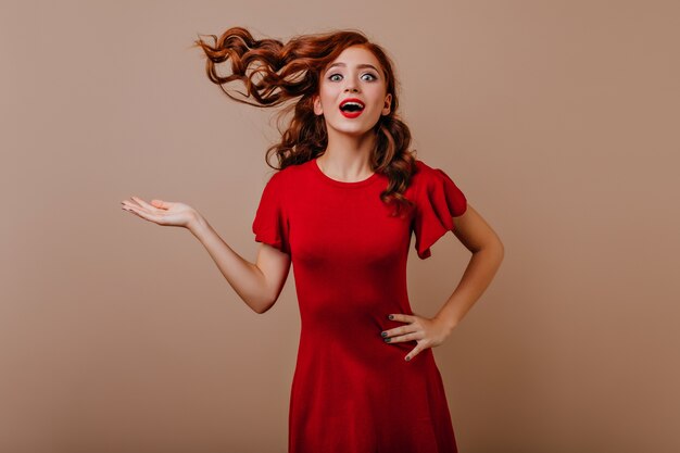 Mujer bonita sorprendida posando juguetonamente. Chica blanca positiva en vestido rojo que muestra verdaderas emociones.