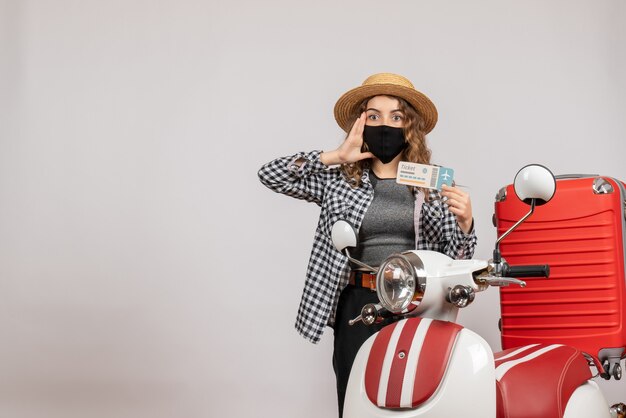 Mujer bonita sorprendida que sostiene el boleto de pie cerca de la maleta roja del ciclomotor