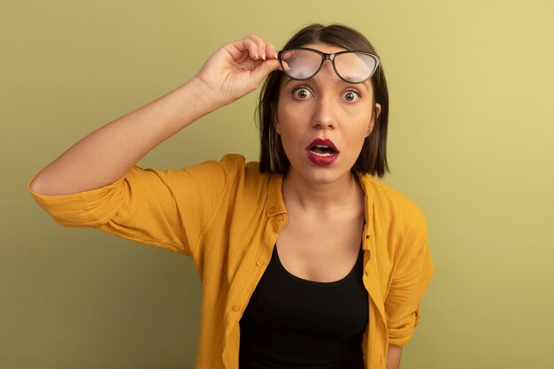 Mujer bonita sorprendida sostiene gafas ópticas y mira al frente aislado en la pared verde oliva