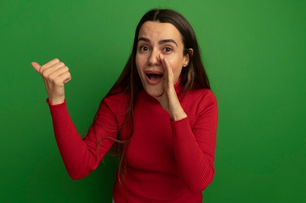 Mujer bonita sorprendida sostiene la mano cerca de la boca y apunta al lado aislado en la pared verde