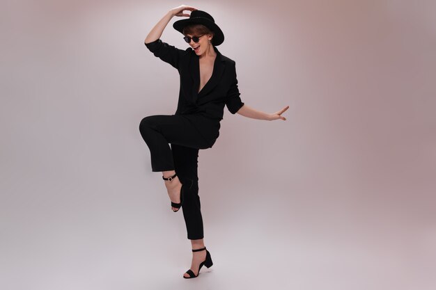 Mujer bonita en traje negro y sombrero se mueve sobre fondo blanco. Encantadora dama en chaqueta oscura y pantalones baila y salta en aislados