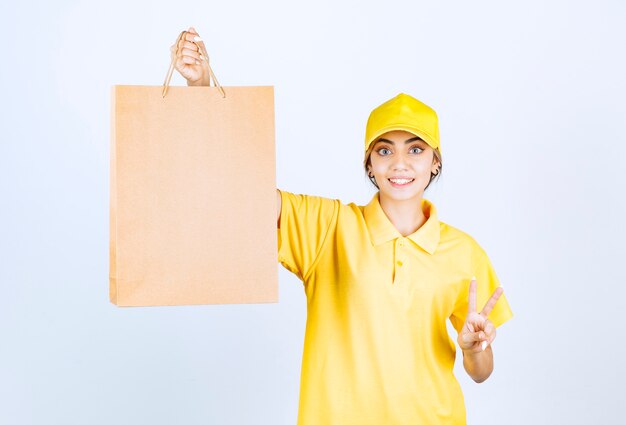 Una mujer bonita en uniforme amarillo con una bolsa de papel artesanal en blanco marrón que muestra el signo de la victoria.