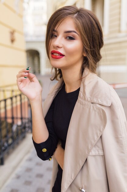 Mujer bonita vestida con vestido negro y trinchera beige con peinado elegante y labios rojos en la calle