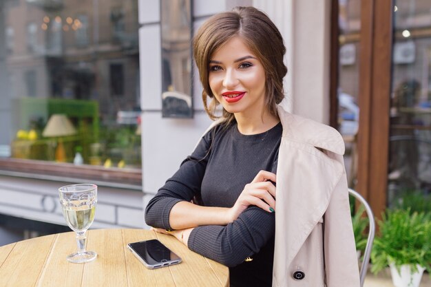 Mujer bonita vestida con vestido negro y trinchera beige con peinado elegante y labios rojos en una terraza