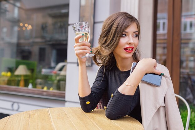 Mujer bonita vestida con vestido negro y trinchera beige con peinado elegante y labios rojos en una terraza