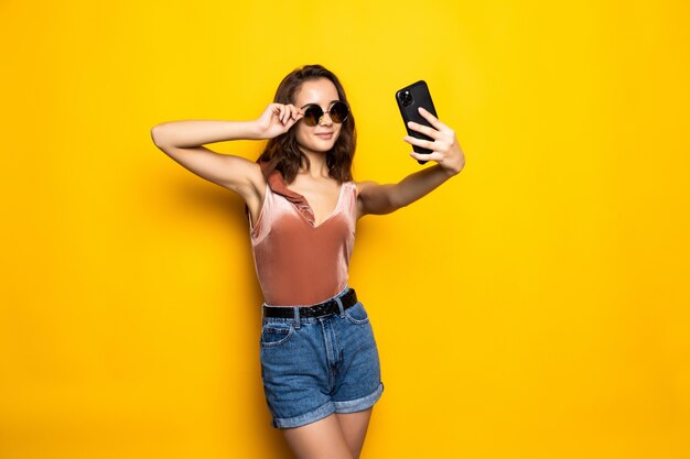 Mujer bonita en vestido haciendo selfie aislado sobre amarillo