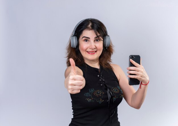 Mujer bonita vistiendo blusa negra mirando haciendo feliz pulgar hacia arriba en auriculares sosteniendo teléfono