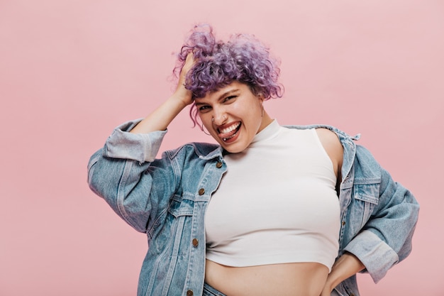 Mujer brillante traviesa con pelo corto morado y sonrisa blanca como la nieve en chaqueta de mezclilla holgada divirtiéndose.