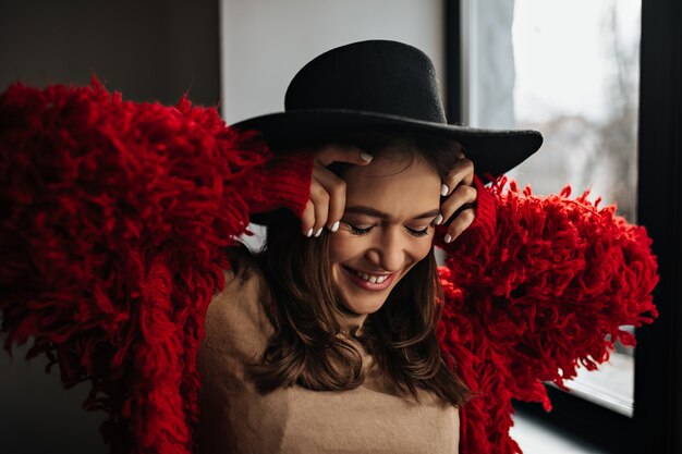 Mujer bronceada de buen humor está posando junto a la ventana. Foto de dama en suéter de punto rojo y sombrero negro.