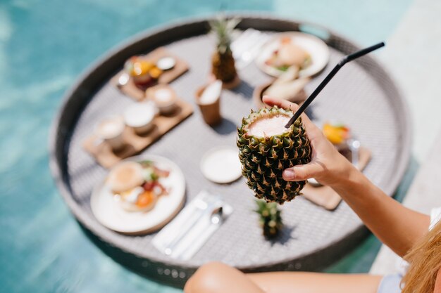 Mujer bronceada con cóctel de piña dulce. Modelo femenino posando durante el almuerzo en la piscina.