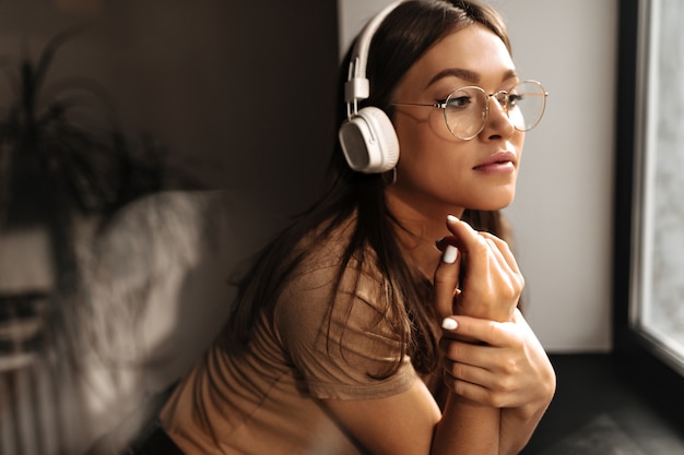 Mujer bronceada con gafas mira por la ventana. Mujer en camiseta beige escuchando canciones en auriculares.