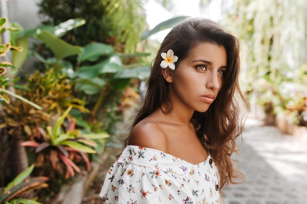 Mujer bronceada pensativa en blusa blanca posando en el jardín