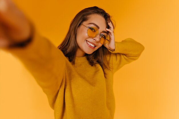 Mujer bronceada refinada con hermosa sonrisa haciendo selfie. Filmación en interiores de una atractiva chica morena en suéter de otoño.