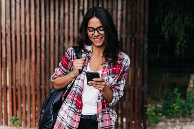 Mujer bronceada romántica de pie al aire libre con mochila y smartphone. Chica latina alegre en gafas lee el mensaje de teléfono con una sonrisa.