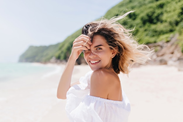 Mujer bronceada soñadora mirando por encima del hombro mientras está de pie en la costa del mar. Adorable mujer rubia posando con placer en la isla tropical en un día caluroso.