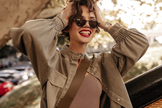 Mujer de buen humor con cabello moreno y labios rojos riendo afuera. Mujer moderna en chaqueta vaquera y gafas sonriendo al aire libre.