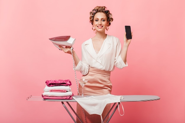 Mujer de buen humor posa con smartphone y plancha cerca de la tabla de planchar