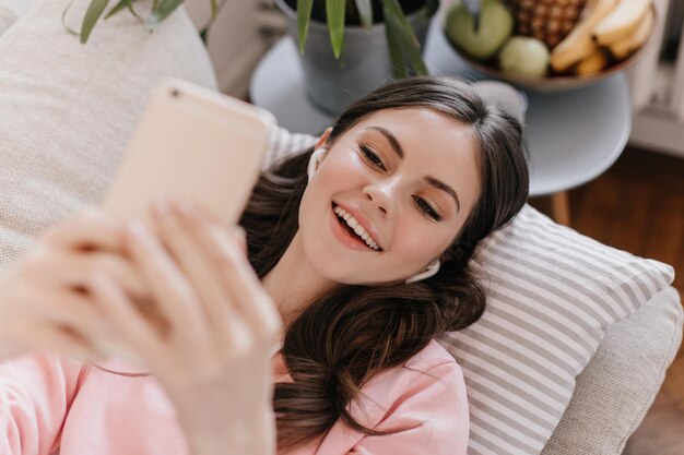 Mujer de buen humor se toma selfie en la sala de estar Chica escuchando música y acostada en el sofá en casa Mujer atractiva sonriendo y sosteniendo un teléfono inteligente