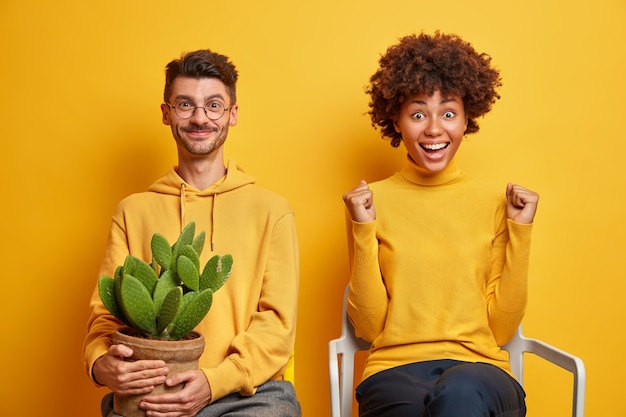 Mujer con cabello afro aprieta los puños se siente muy contento celebra el éxito posa en una cómoda silla cerca del novio aislado en amarillo