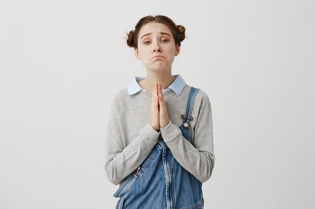 Mujer con cabello castaño en doble bollos posando con piedad mirada tomados de la mano en la oración Patéticas emociones de niña pidiendo perdón sobre la pared blanca. Concepto de emociones