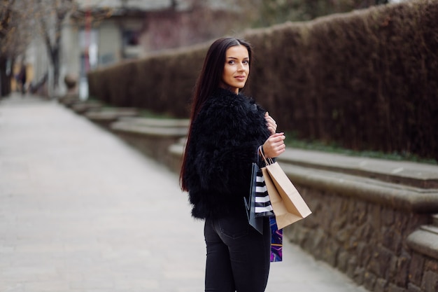 Una mujer de cabello castaño vestida de negro, sostiene bolsas de compras coloridas y estampadas durante una exitosa juerga de compras. Caminando afuera, ella disfruta del calor de un día.