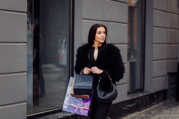 Una mujer de cabello castaño vestida de negro, sostiene bolsas de compras coloridas y estampadas durante una exitosa juerga de compras. Caminando afuera, ella disfruta del calor de un día.