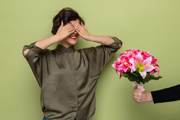 Mujer con cabello corto mirando sorprendida cubriéndose los ojos con las manos mientras recibe un ramo de flores de su novio celebrando el día internacional de la mujer el 8 de marzo de pie sobre fondo verde