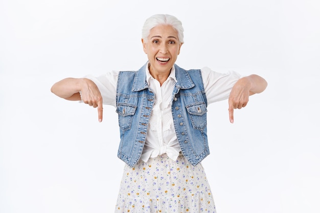 mujer con cabello gris peinado, usar chaleco de mezclilla, vestirse, señalar con el dedo hacia abajo, sonreír y reír divertida, mirar cámara interesada y emocionada