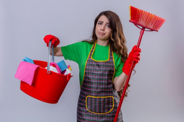 Mujer con cabello largo ondulado vistiendo delantal y guantes de goma sosteniendo un balde con herramientas de limpieza y un trapeador con aspecto triste y trabajado con cara infeliz de pie