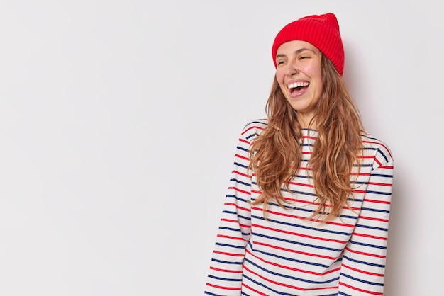 mujer con cabello largo sonríe sinceramente se siente muy feliz viste un jersey a rayas casual y un sombrero rojo estando de buen humor posa contra el espacio de copia en blanco blanco para su anuncio