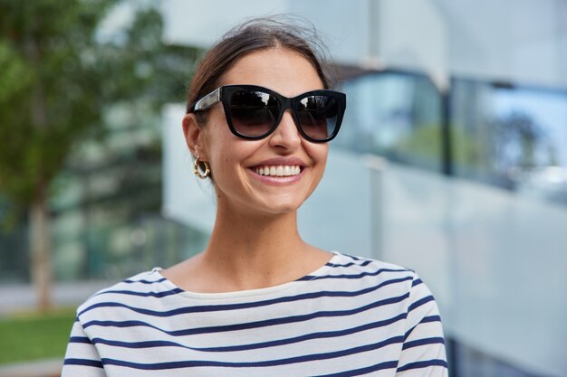 Mujer con cabello oscuro chilld durante el bonito día de primavera lleva gafas de sol puente de rayas plantea en borrosa