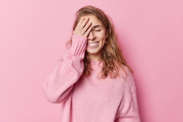 Una mujer de cabello oscuro complacida hace que la palma de la cara trate de esconderse, mantiene los ojos cerrados, sonríe suavemente y se ríe de algo que usa un puente casual aislado sobre un fondo rosado. Toma monocromática. Concepto ciego.