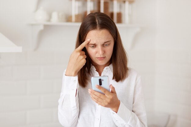 Mujer de cabello oscuro confundida y perpleja de pie con el teléfono celular en las manos, revisando las redes sociales, leyendo un mensaje o comentario negativo, posando en la cocina de su casa.