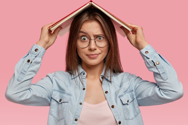 La mujer de cabello oscuro descontenta se ve con expresión insatisfecha, se siente cansada del aprendizaje constante, mantiene el libro sobre la cabeza, usa camisa de mezclilla, gafas ópticas, exige vacaciones, aislado sobre una pared rosa