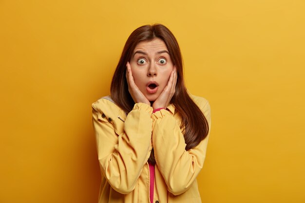 Mujer de cabello oscuro emocional asustada mira fijamente con ojos saltones, mantiene la boca abierta, aprendió algo aterrador, usa chaqueta, aislada en la pared amarilla. Concepto de reacción humana