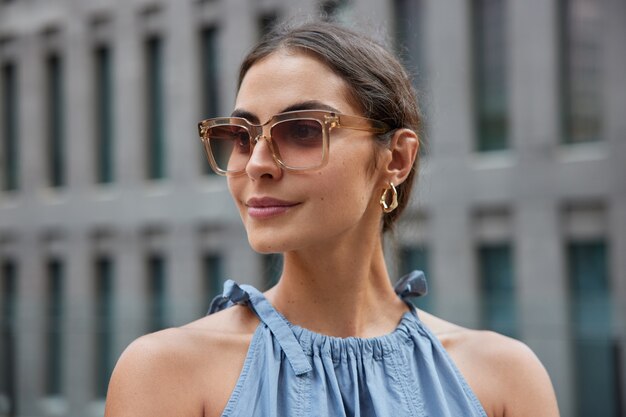 Mujer con cabello oscuro enfocado con expresión satisfecha lleva gafas de sol de moda y camina por la ciudad discons algo nuevo plantea en borrosa