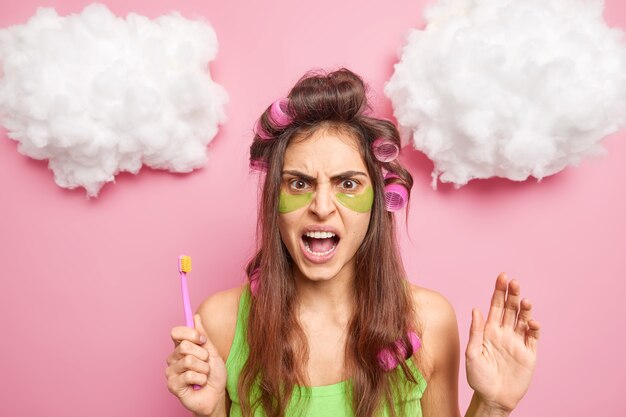 Mujer de cabello oscuro enojada disgustada aplica parches de colágeno rizadores de pelo debajo de los ojos para reducir las arrugas y las líneas finas sostiene el cepillo de dientes tiene rutinas de higiene bucal diarias aisladas en la pared rosa del estudio