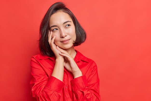 mujer con cabello oscuro mantiene las manos cerca de la cara tiene expresión pensativa imagina algo en mente viste camisa roja en un tono con