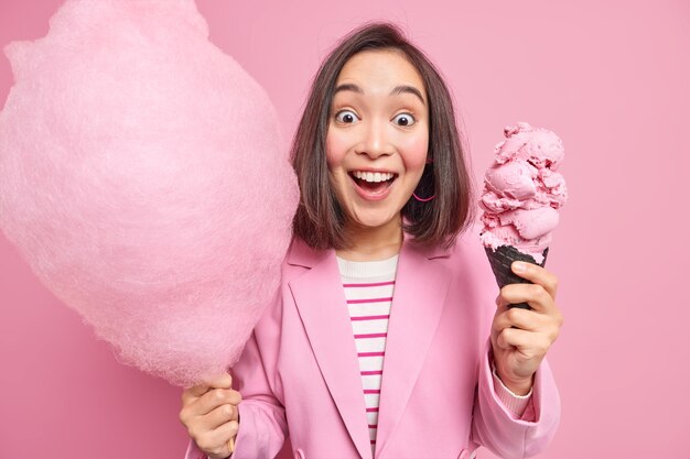 Mujer con cabello oscuro mira impresionado a la cámara sostiene algodón de azúcar y sabroso helado apetitoso vestido formalmente