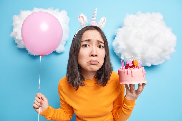 Mujer con cabello oscuro mira tristemente a la cámara celebra el cumpleaños solo tiene un delicioso pastel y un globo inflado aislado en azul